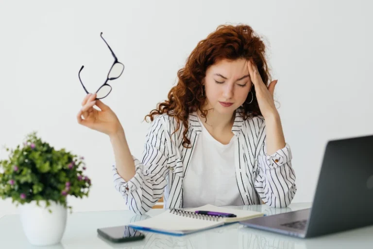 Woman at her desk holding her glasses with a hand to her head, looking overwhelmed